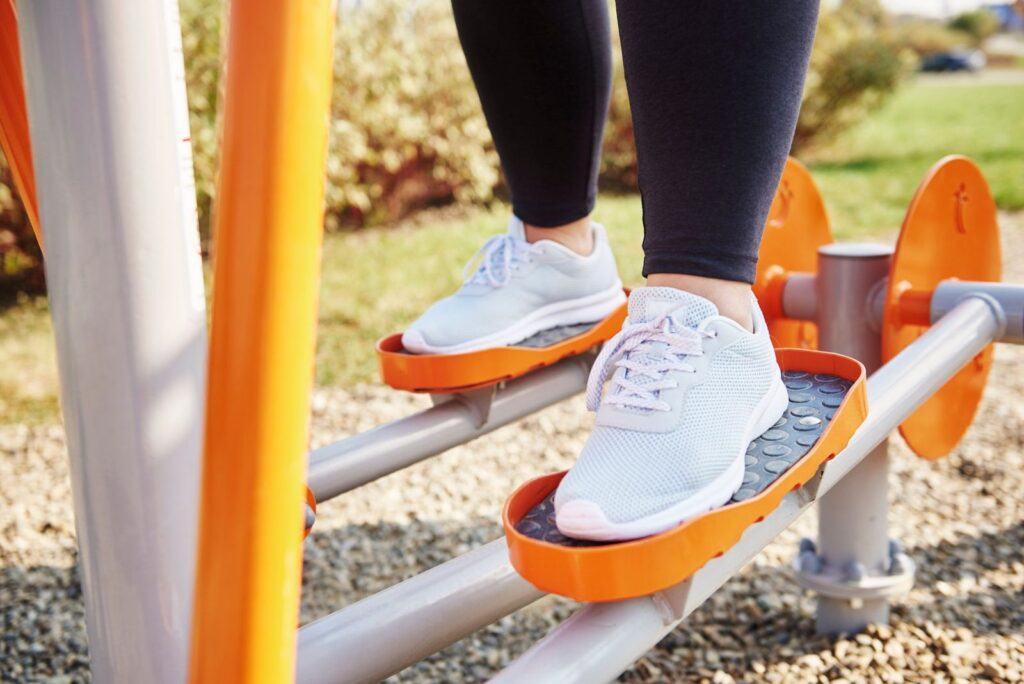 Feet in white sneakers on the orange footplates of an outdoor elliptical machine, showing low-impact plantar fasciitis exercises in a park